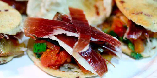 Una tapa con forma de bocadito pequeño y fácil de comer. Masa de pan y trocitos de jamón, un aliño de tomate y albahaca para darle jugosidad al pan y coronado por una lámina de Boletus Edulis confitado y jamón ibérico de Bellota Cinco Jotas.